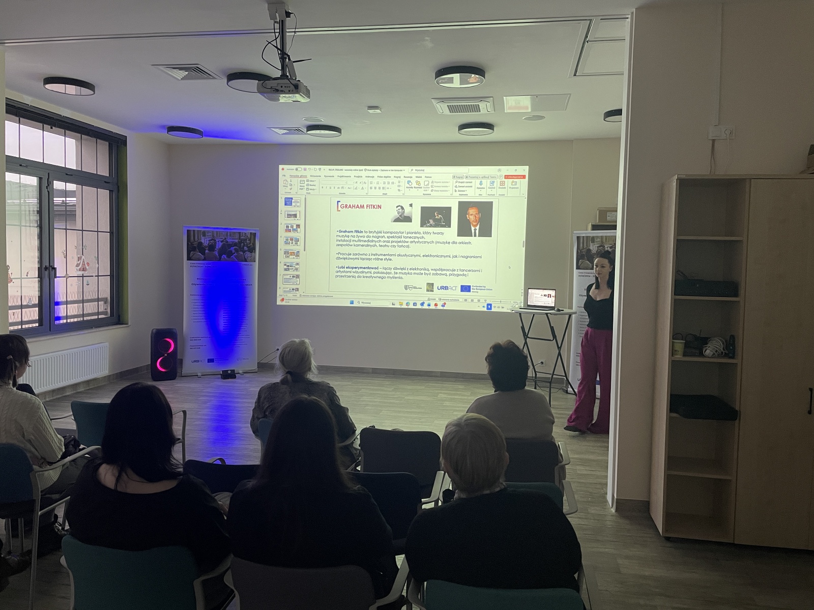 Participants during a presentation in the room – looking at a slide on the screen showing the biography of British composer Graham Fitkin.