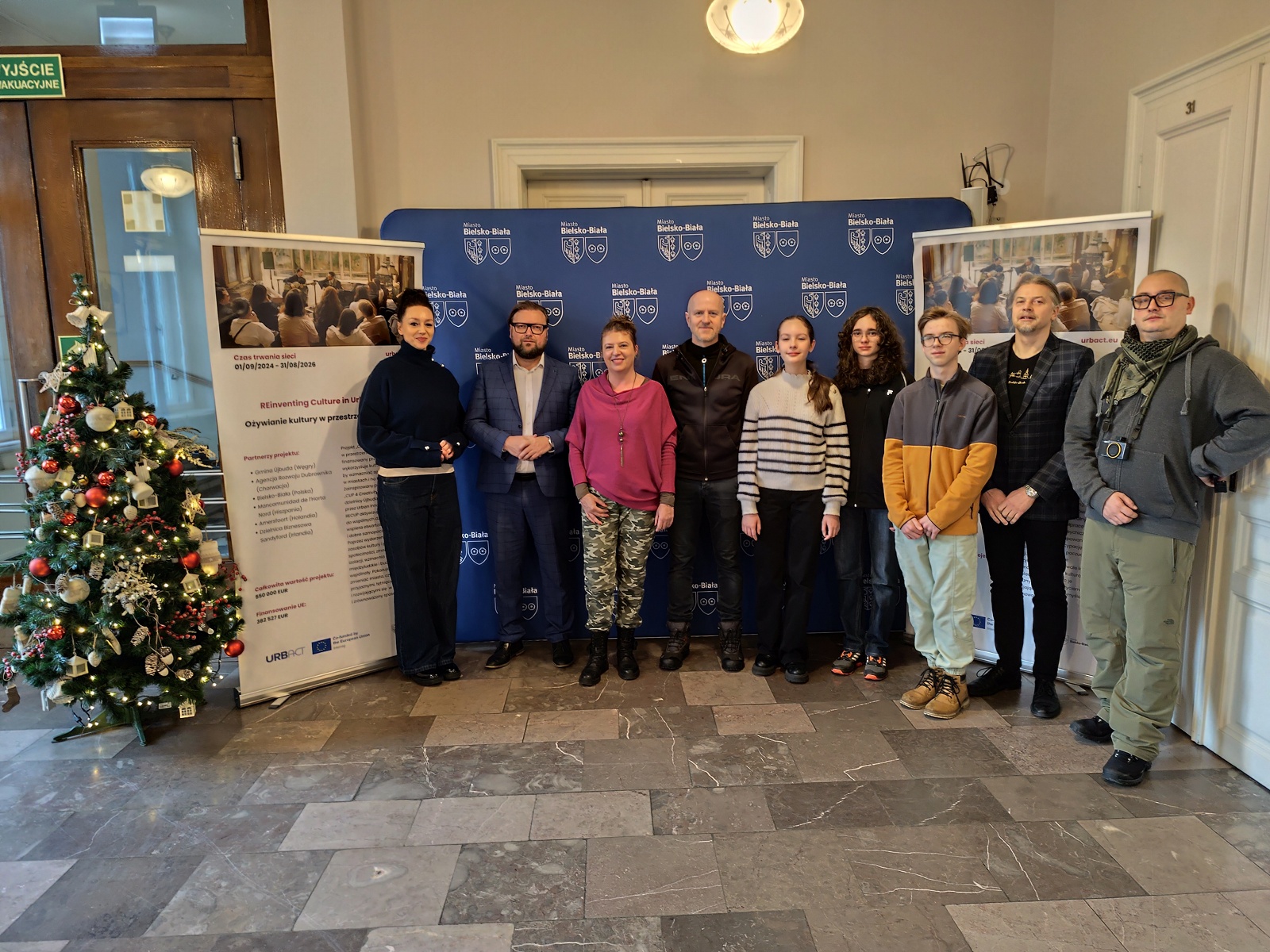 Group of participants in a joint photograph with representatives of the city authorities and municipal office, inside a building – standing in front of banners and a Christmas tree.