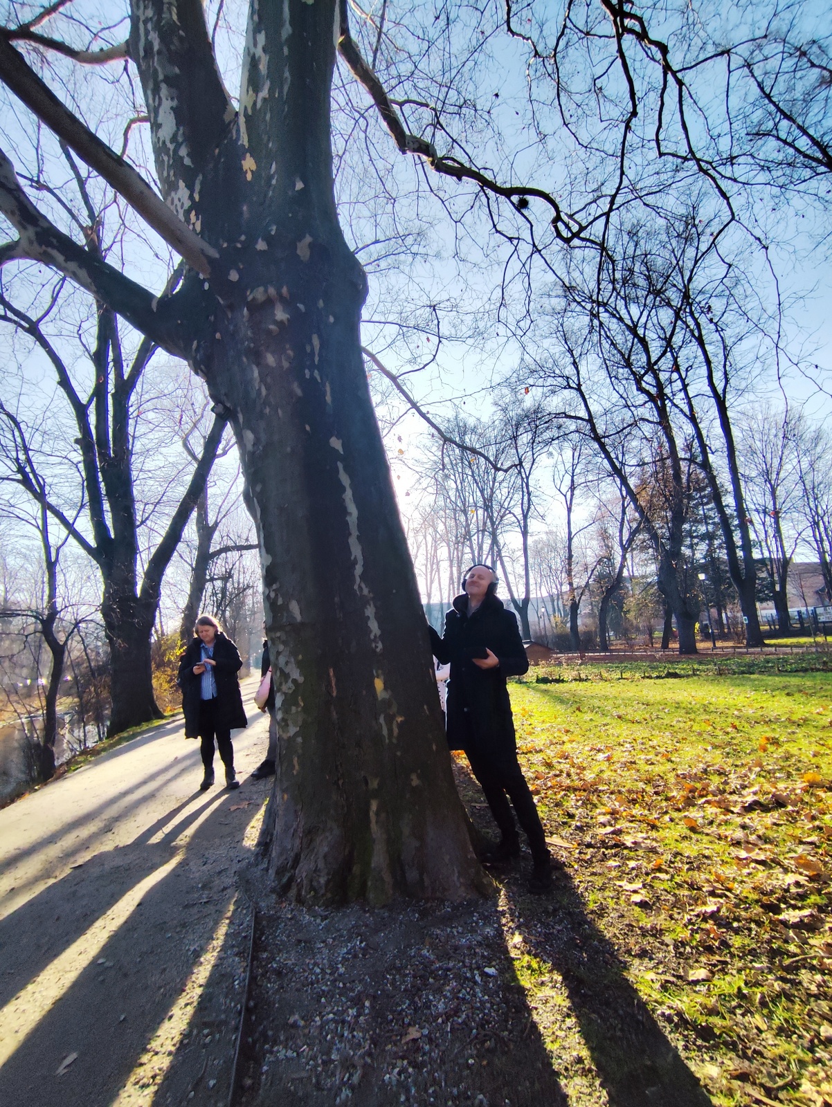 Local musician standing beside a tree with sound-recording equipment and headphones, eyes closed and smiling, listening to sounds from inside the tree.