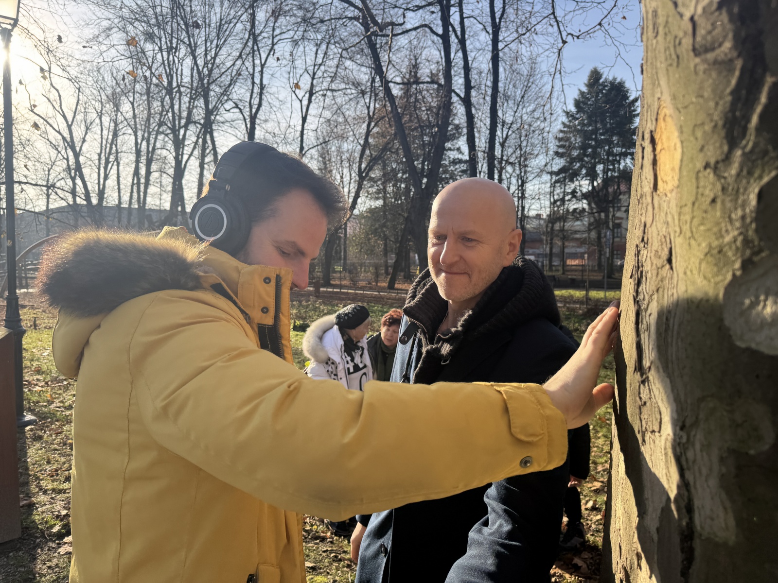 Two people beside a tree. One, wearing headphones, touches the bark with a hand while listening to sounds from inside the tree.