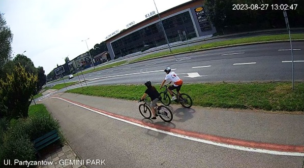 zdjęcie przedstawiające dwóch rowerzystów na ścieżce rowerowej, biegnącej zdróż drogi w otoczeniu budynków i zieleni 