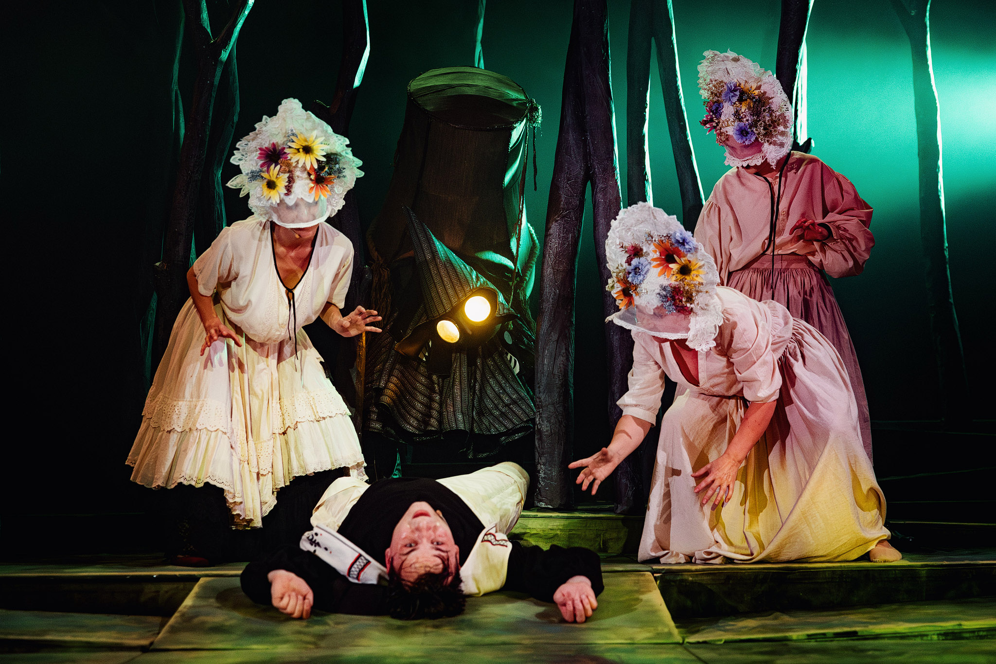 Na zdjęciu trzy postaci kobiece w kapeluszach z kwiatami pochylają się nad chlopcem