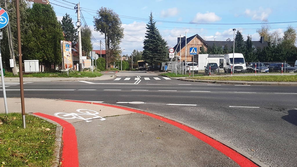 zdjęcie przedstawiające fragment drogi, znaki drogowe, budynki, zaparkowane pojazdy oraz zieleń