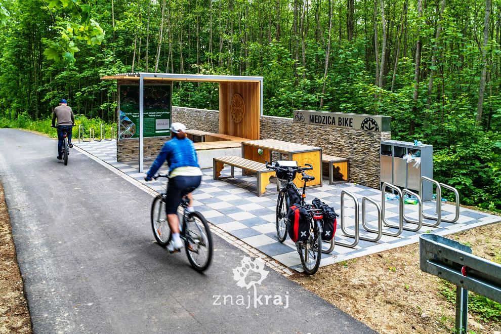 zdjęcie przedstawiające rowerzystów na ścieżce rowerowej, ławeczki, zadaszenie, stojaki na rowery na tle lasu