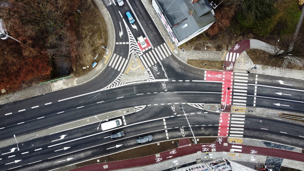 grafika przedstawiająca zdjęcie z lotu ptaka przedstawiające fragment ulicy z przejściami dla pieszych, śluzami dla rowerów, chodnikami oraz pojazdami