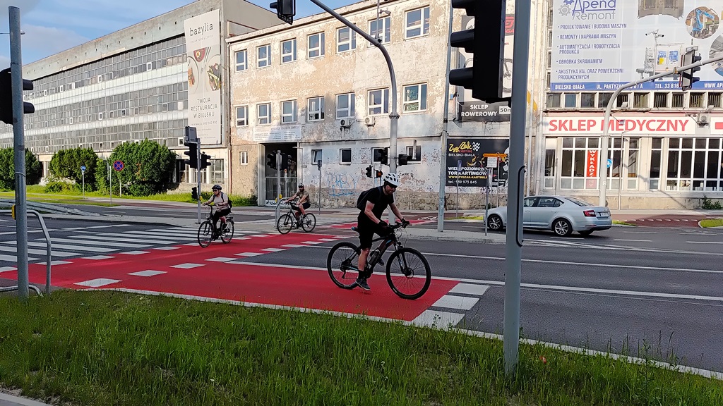 grafika przedstawiająca zdjęcie fragmentu ulicy z przejściem dla pieszych, śluzą dla rowerów, rowerzystami, pojazdem oraz w tle budynki oraz zieleń