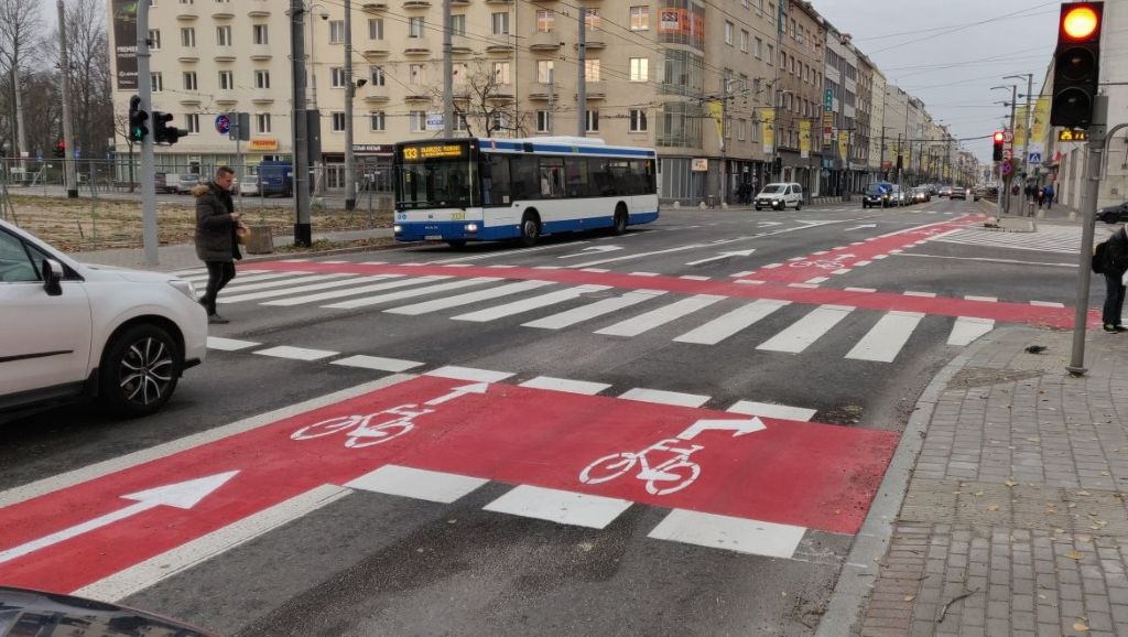 zdjęcie przedstawiające śluzę dla rowerów, jezdnię, pieszego, pojazdy, światła drogowe oraz budynki