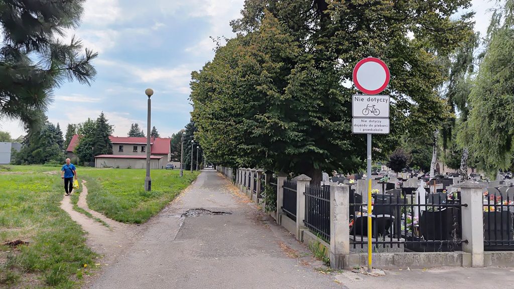 zdjęcie przedstawiające drogę obok cmentarza, znaki drogowe, przechodnia, budynki oraz zieleń