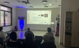 Participants during a presentation in the room – looking at a slide on the screen showing the biography of British composer Graham Fitkin.