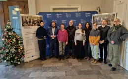Group of participants in a joint photograph with representatives of the city authorities and municipal office, inside a building – standing in front of banners and a Christmas tree.