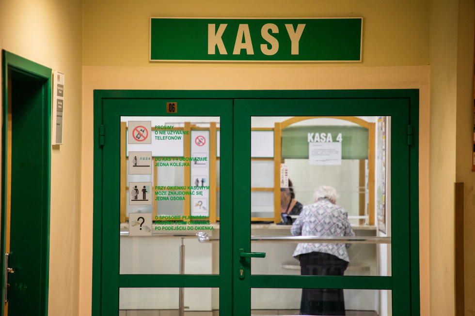Na zdjęciu osoba stojąca przed okienkiem kasowym, nad drzwiami napis KASY
