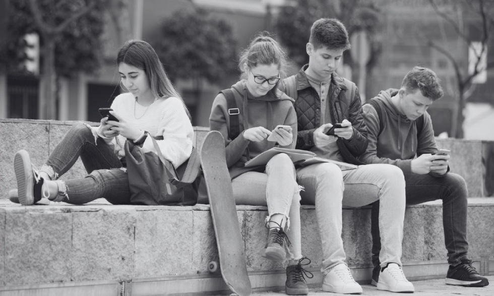 Na zdjęciu młodzież na zewnątrz, siedzi na murku z telefonami w rękach 