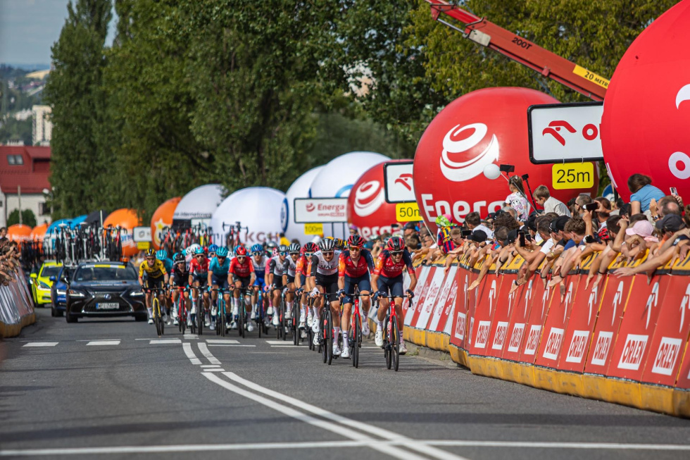 Na zdjęciu peleton kolarzy na drodze, kibice za ogrodzeniem
