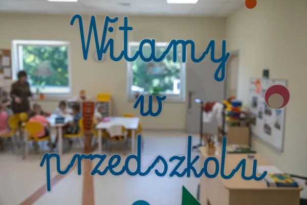 Na zdjęciu napis Witamy w przedszkolu na szybie, przez którą widać salę przedszkolną